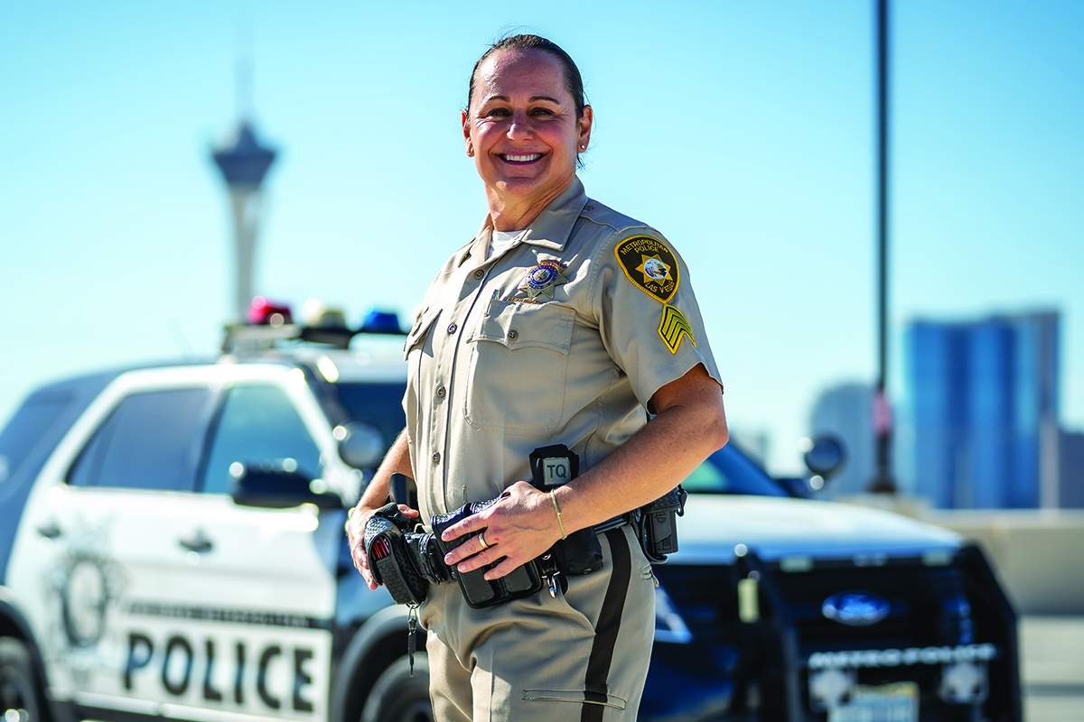 A woman in a police uniform stands smiling in front of a police car.