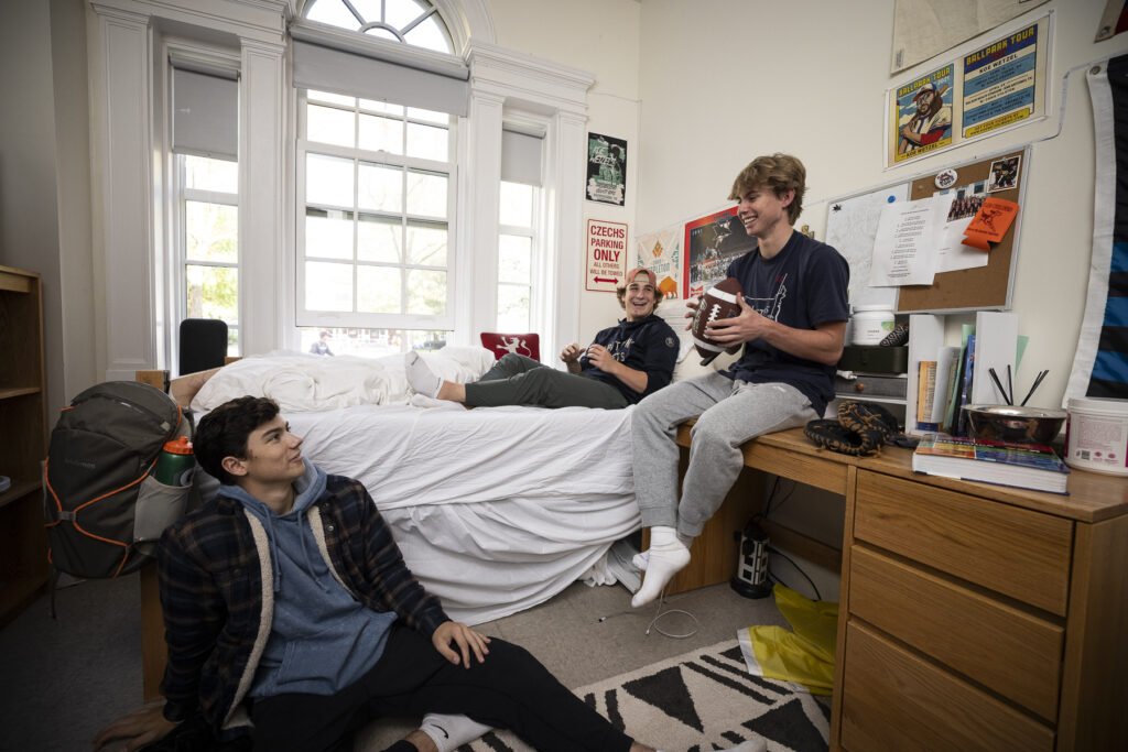 Three boys sit around dorm room.