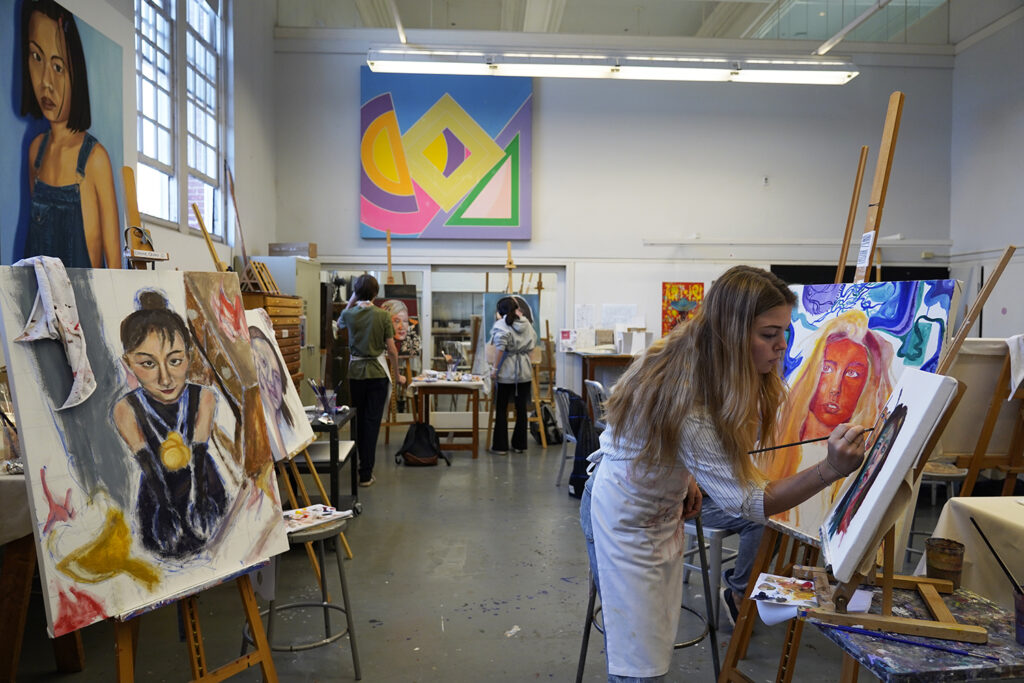 Student surrounded by canvases painting self portrait