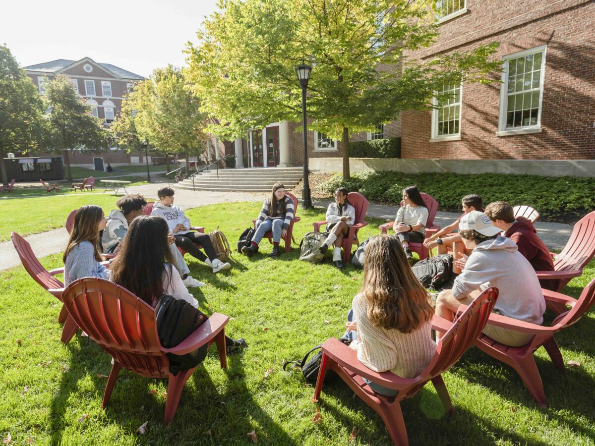 Group of students sit on red chairs outside on a green quad lawn