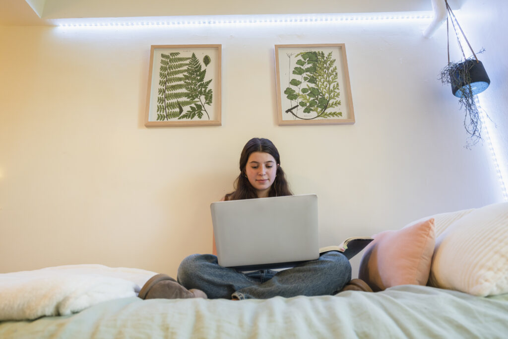 Girl sits on bed in dorm room on laptop