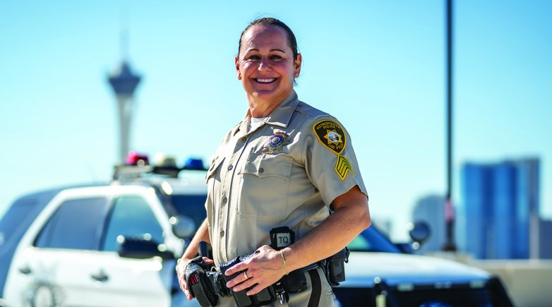 A woman in a police uniform stands smiling in front of a police car.