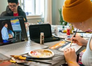 Student wearing a beanie paints while Bob Ross video plays on computer