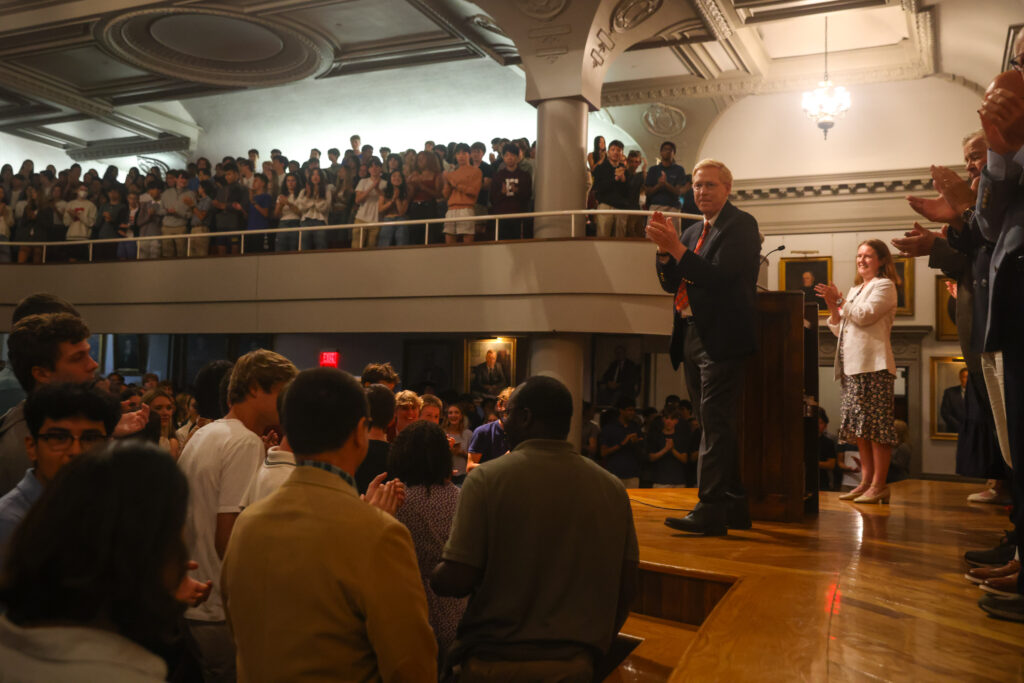 Principal Rawson stands on Assembly Hall stage clapping surrounded by an Assembly Hall full of students