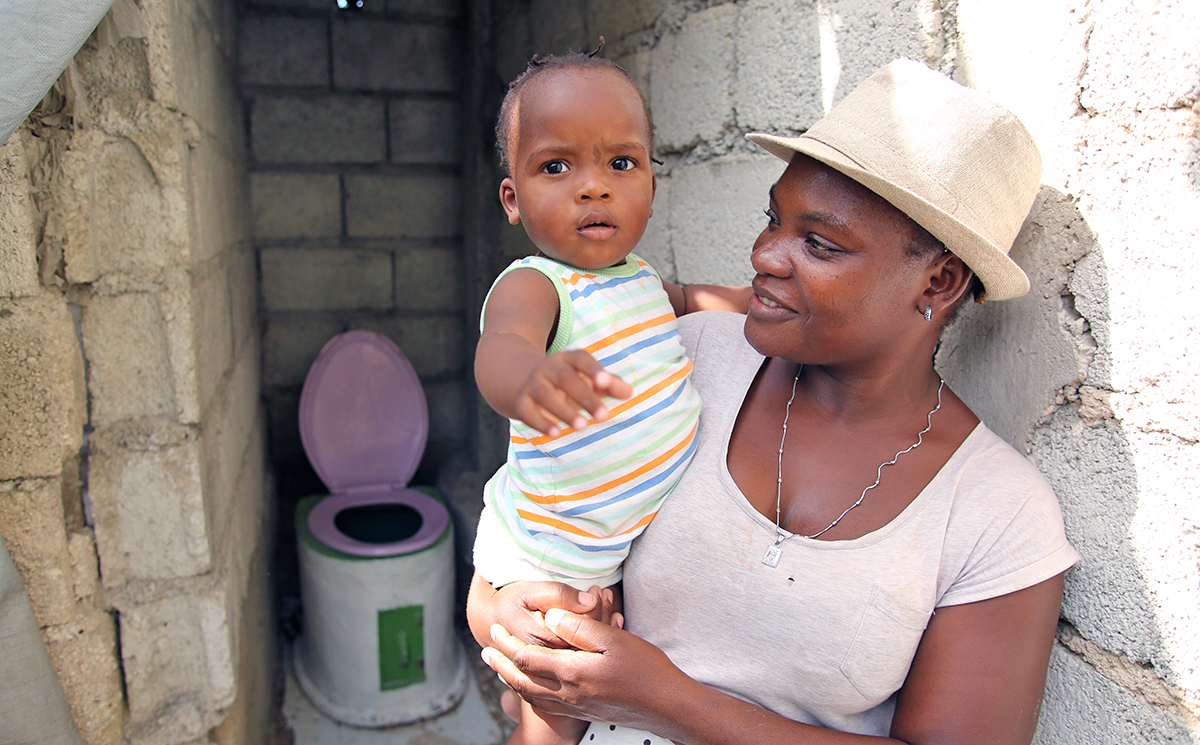Woman holding a child with a SOIL toilet in the background.
