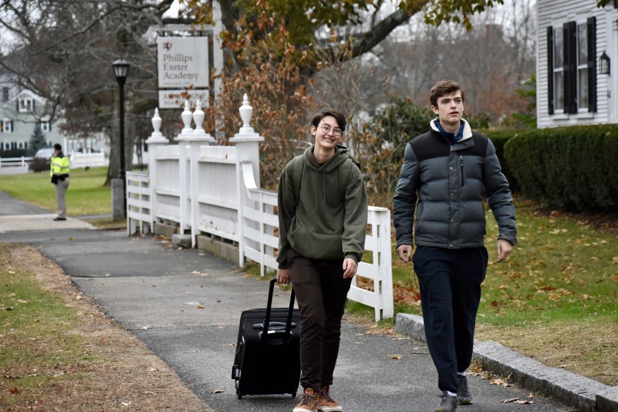 Two students walk down a sidewalk on Exeter's campus, one pulling a suitcase.