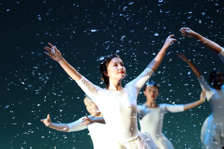 Three students dance dressed in white costumes with snow falling around them. 