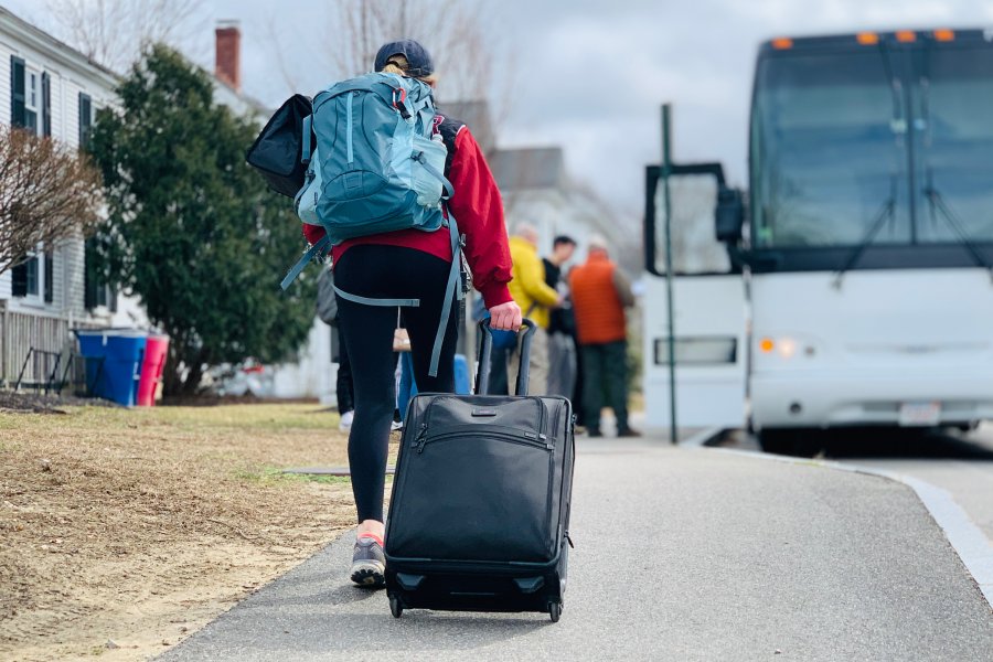 Student wheels a suitcase along the street toward a waiting bus. 