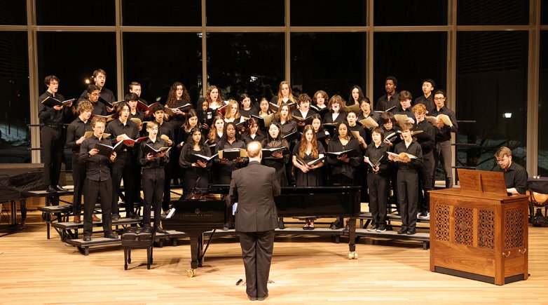 A chorus of students dressed in black standing on risers in front of tall windows at night