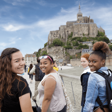 Three female students smiling back over their shoulders in front of Mont-Saint Michel